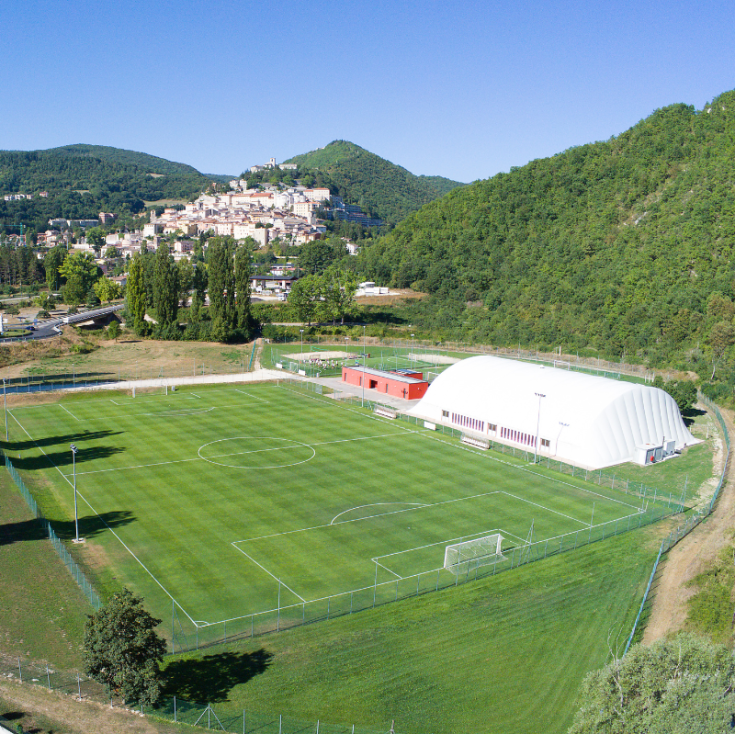 Un campo da calcio situato tra boschi e colline, con una semplice struttura sportiva. Pannelli segnaletici e porte sono presenti.