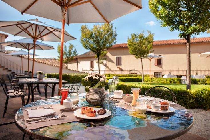 An outdoor breakfast table with various dishes and a view of a well-kept garden.