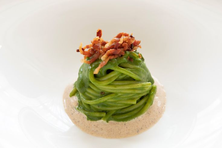 A serving of green pasta topped with crispy pancetta, artfully arranged.