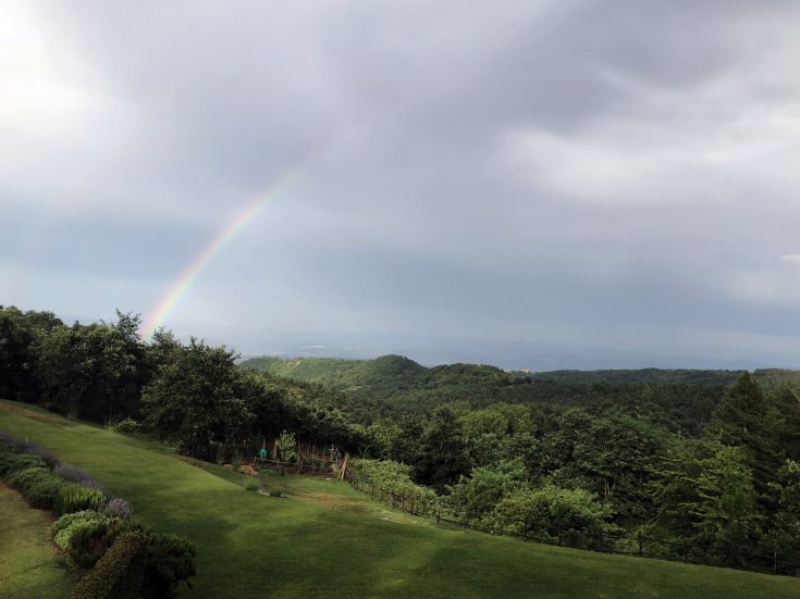 A colorful rainbow stretches over the hills, between clouds and mountains, creating a serene and natural panorama.