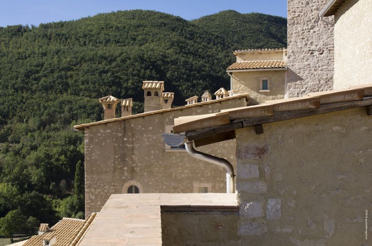 A panoramic view of the medieval village of Postignano, nestled among gentle hills with mountains in the background.