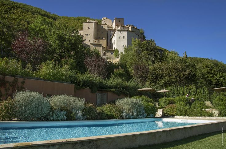 A medieval village surrounded by greenery, featuring a swimming pool and calm scenes of the surrounding landscape.