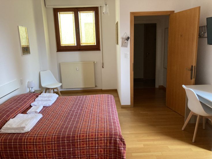 Komfortables Zimmer mit Doppelbett und einfachen Möbeln. Beleuchtet durch große Fenster.