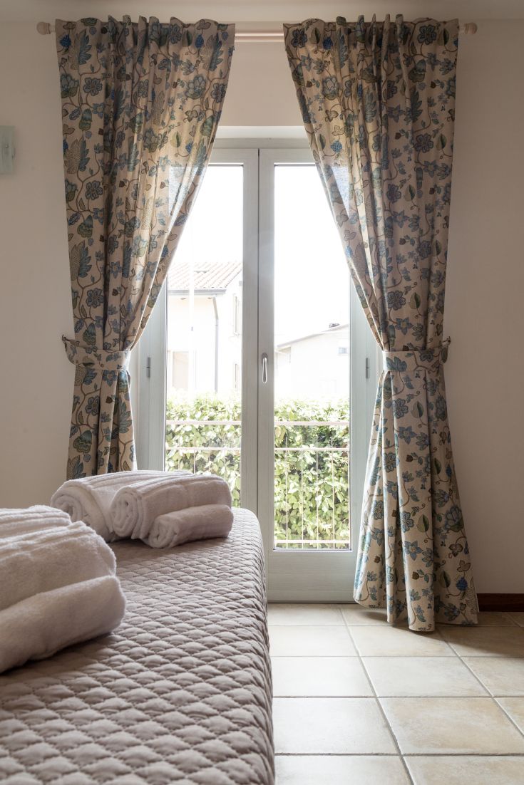 Camera semplice con lenzuola piegate e tende a fiori, affacciata su un giardino.