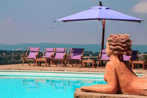 Eine Skulptur einer weiblichen Figur neben einem Pool, mit Liegestühlen und einem blauen Sonnenschirm im Hintergrund.