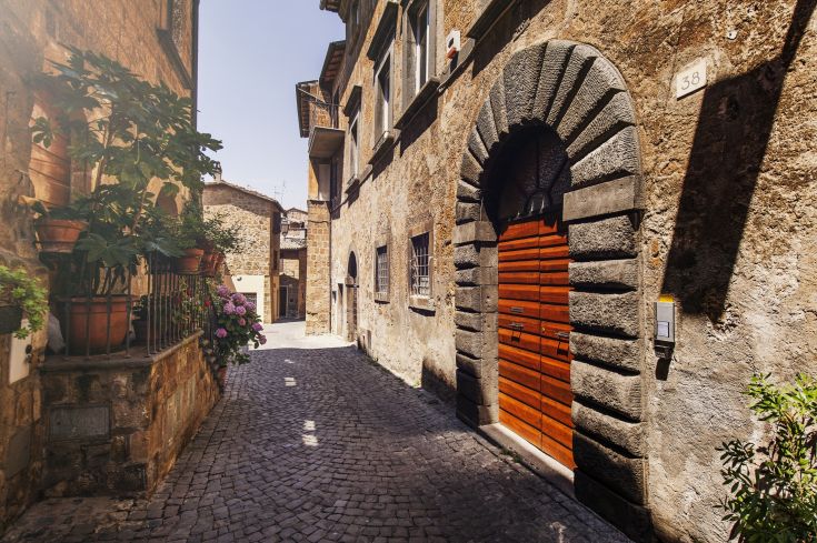 Una stretta strada di un borgo storico, circondata da piante fiorite e caratterizzata da edifici antichi.