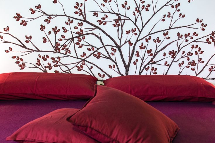 Particolare di un letto con cuscini rossi; la parete presenta decorazioni con rami e bacche rosse.