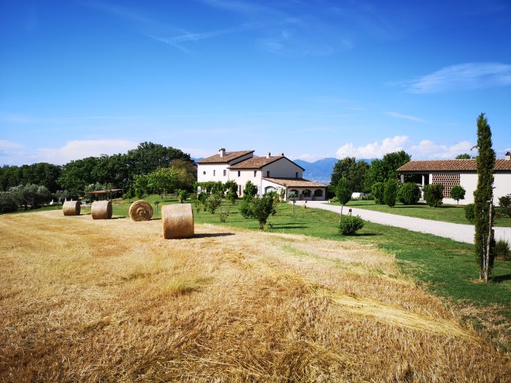 Un casale in campagna immerso nel verde, circondato da prati e balle di fieno sotto un cielo sereno.