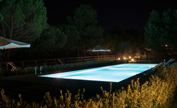Ein Schwimmbecken eines Agriturismo ist nachts beleuchtet und von üppigem Grün umgeben.