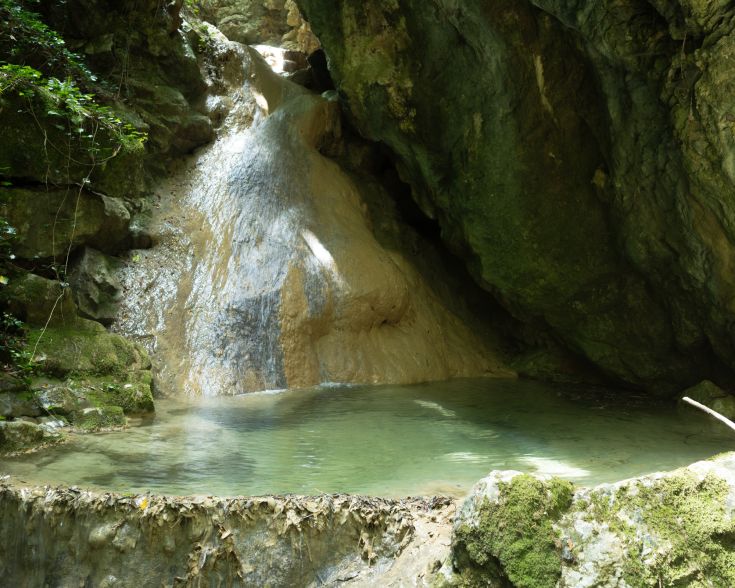 Eine ruhige Szene mit einem Wasserfall, der in einen ruhigen See fließt, umgeben von Felsen und üppigem Grün.