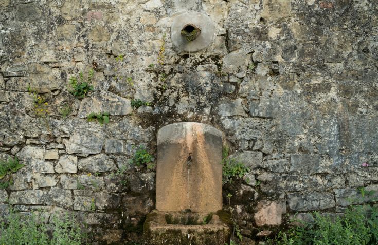 Ein alter Steinbrunnen an einer Wand, umgeben von üppigem Grün. Ein ruhiger und einladender Ort.