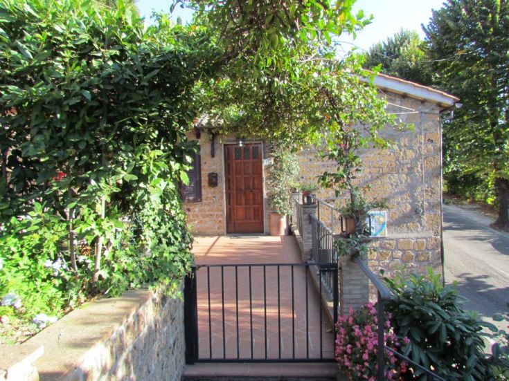 Ingresso di una casa circondata da piante, con elementi in pietra e legno sotto una struttura di pergola.