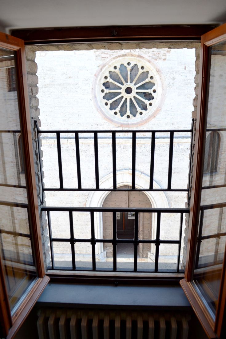 A view from a window with bars, showcasing a historic building. A decorated rose window is visible on a light wall.
