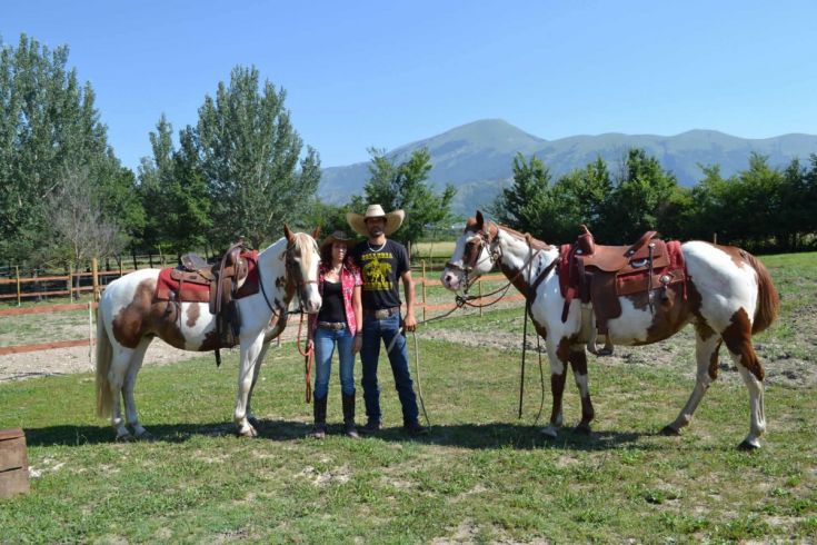 A couple riding a horse in a green meadow, with trees and mountains in the background.