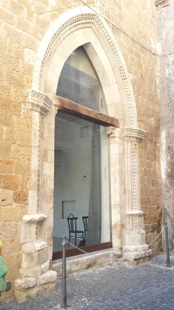 Una porta storica con dettagli architettonici e una finestra contemporanea. L'interno è essenziale, con una sedia semplice.