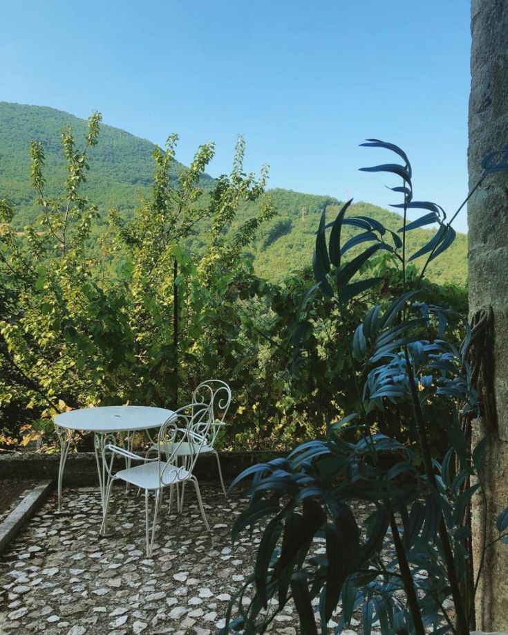 Outdoor area with a table and chair, surrounded by lush greenery and gentle hills.