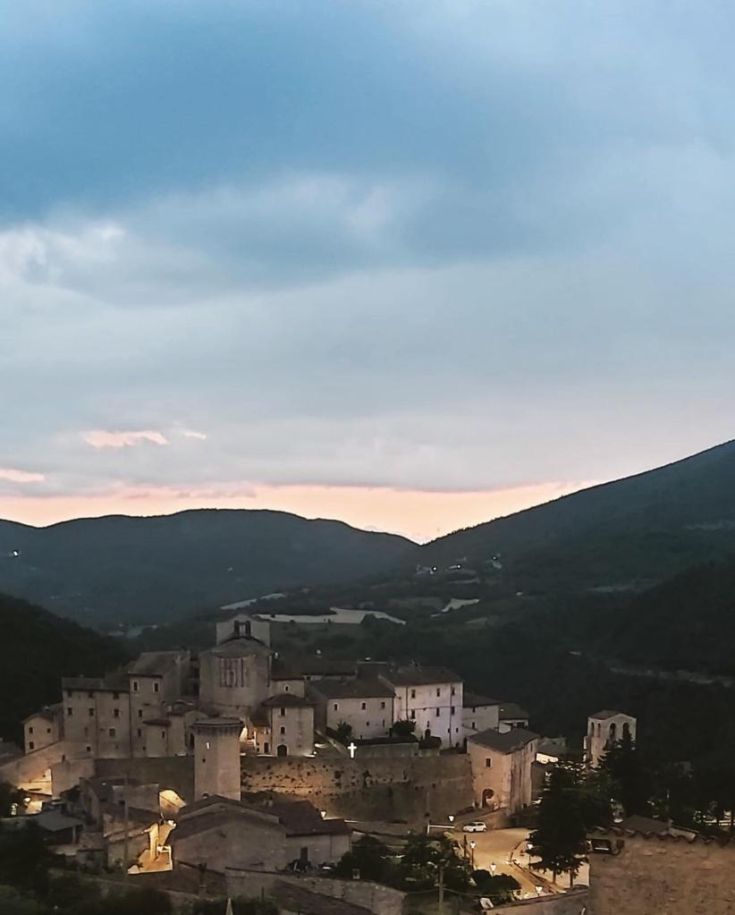 A historic village glows with warm colors at sunset, surrounded by green hills and stone houses.