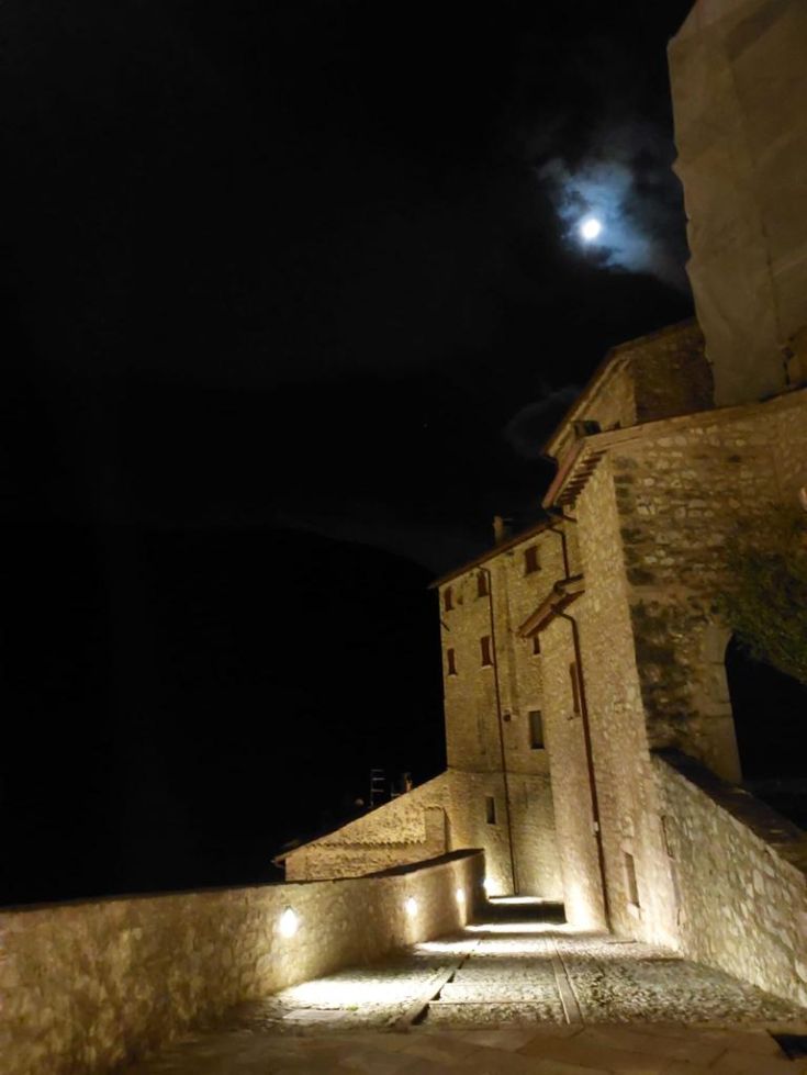 A well-lit stone street winds through a village with historic buildings under a night sky. A serene atmosphere.