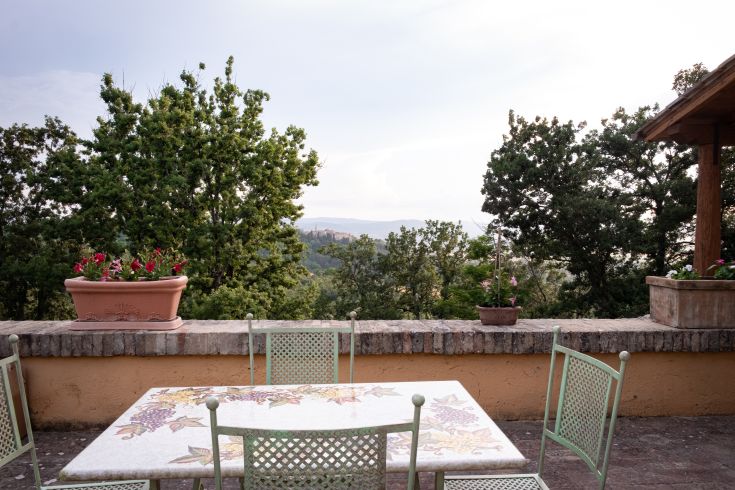 Scène de campagne ombrienne avec une table et des chaises vertes, entourées d'arbres et de plantes fleuries.