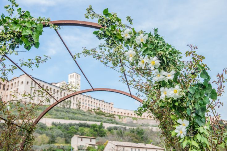 Un arco fiorito che si apre su un antico monastero, immerso nel paesaggio di Assisi.