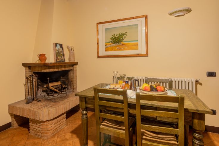 Spazio semplice con un tavolo, frutta di stagione e un camino, in un ambiente di campagna.