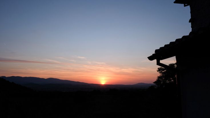 A sunset over a valley, featuring a silhouetted house on the horizon.