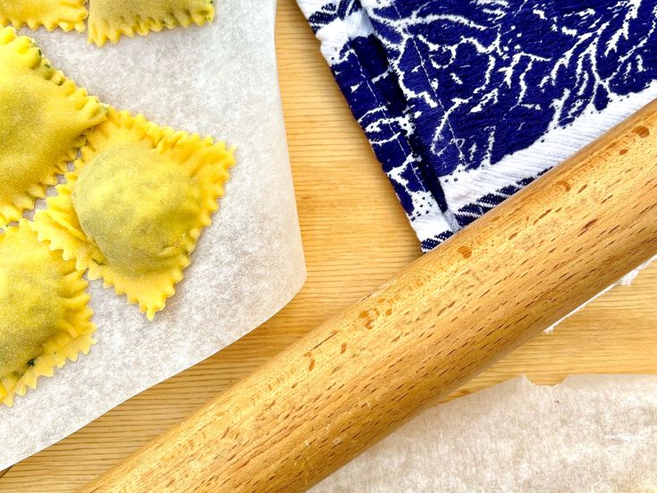 Vorbereitung von frischen Ravioli auf einem hellen Holztisch mit einem Nudelholz und einer karierter Tischdecke.