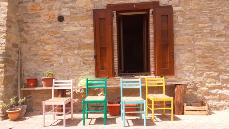 A group of colorful wooden chairs placed in front of a stone building with an open window.