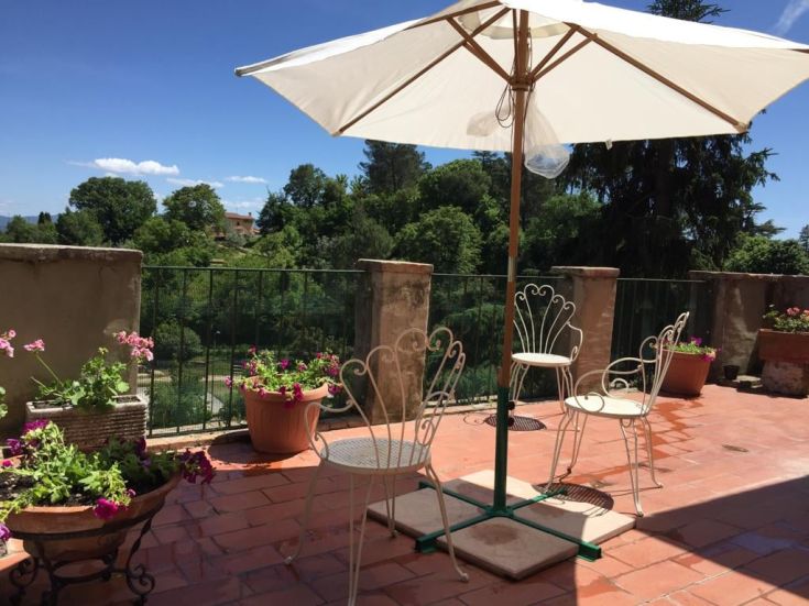 Terrazza con ombrellone e sedie, circondata da piante fiorite, panoramica su un paesaggio verde.