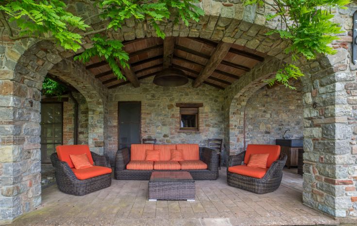 Ein Innenhof eines Gebäudes mit Rattan-Sofa und -Stühlen, ausgestattet mit orangefarbenen Kissen, umgeben von Grün.