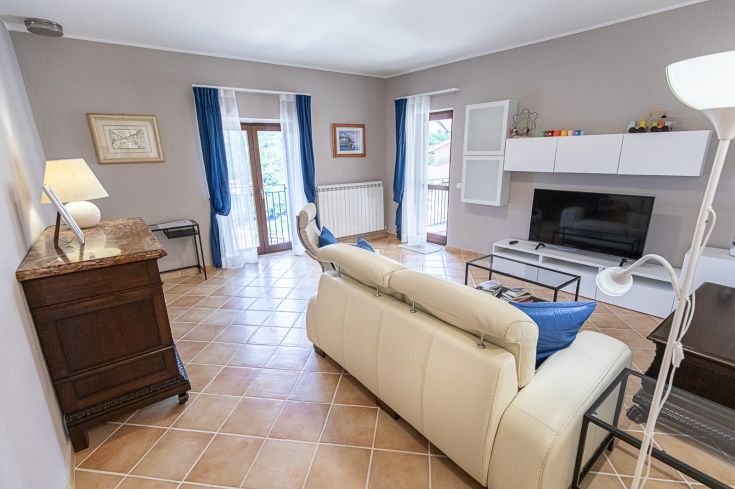 Salon lumineux avec un canapé clair, une télévision et un accès à un jardin en pierres.