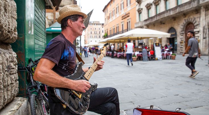 Un musicista suona la chitarra in una piazza affollata, circondato da passanti che si spostano tra i tavoli.