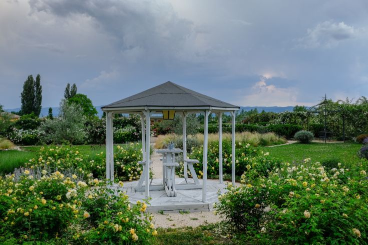 Gazebo bianco situato in un giardino con rose circondanti e vegetazione, in una giornata nuvolosa.