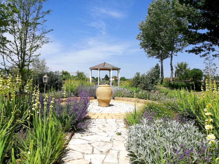 Un giardino ben curato con un gazebo e un vaso centrale, circondato da varie piante e fiori, in un ambiente rilassato.