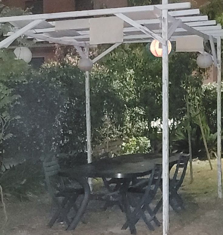 Una pergola semplice con tavoli e sedie in un giardino tranquillo, decorata con lanterne soffuse.