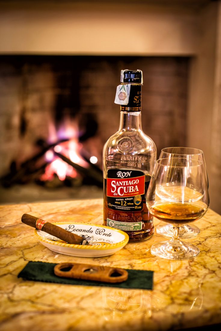 Un bicchiere di rum e un sigaro posati su un tavolo accanto a un camino, in un ambiente accogliente.