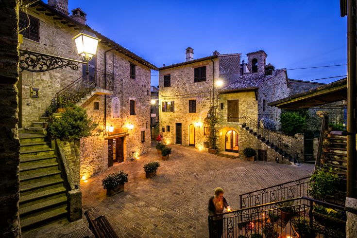 Vista di una piazza medievale illuminata nella serata, con edifici in pietra e un'atmosfera tranquilla.