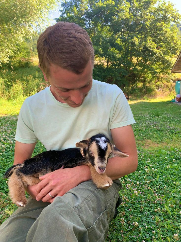 Ein Mann umarmt ein verspieltes Zicklein auf einer grünen Wiese, umgeben von einer friedlichen Atmosphäre.
