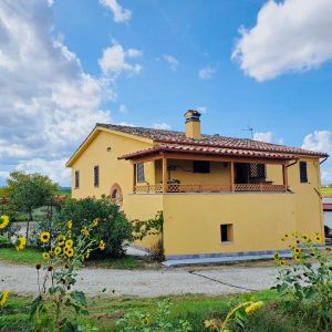 Casa gialla circondata da spazi verdi, con un portico che si affaccia su un giardino con fiori variopinti.
