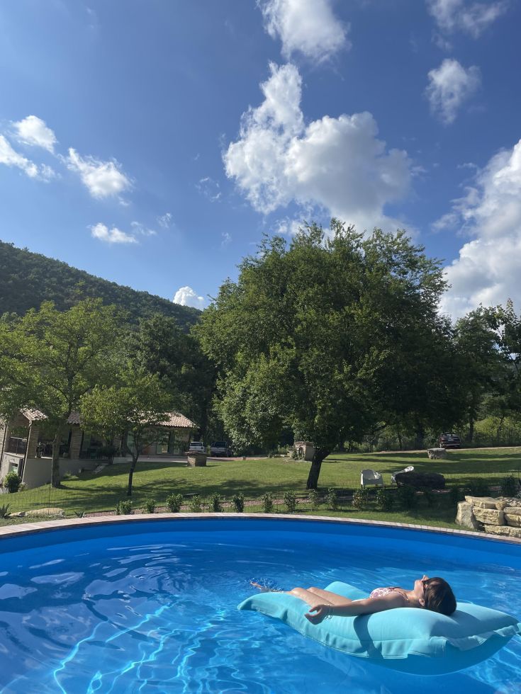A woman lounges on a floating mat in a pool, surrounded by trees under a clear blue sky.