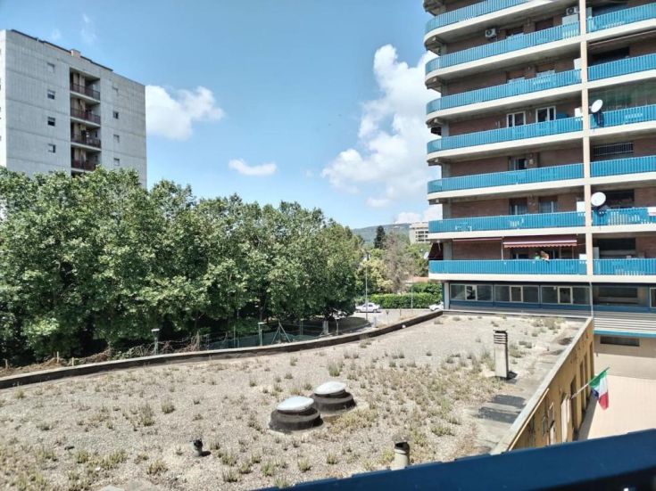 Panorama di un'area residenziale, con alberi e edifici sullo sfondo, cielo sereno e un prato verde.