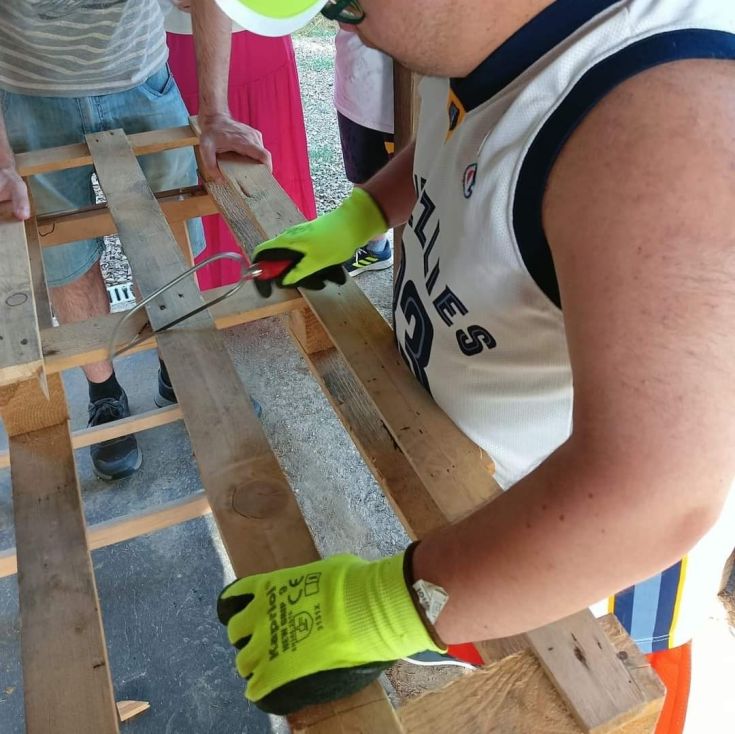 A man works on a wooden bench, using cutting tools in an outdoor setting.