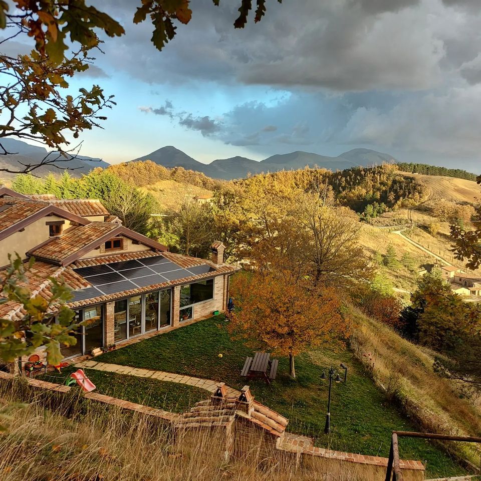 Scena autunnale con un edificio circondato da vegetazione e montagne sullo sfondo.