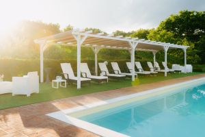 Un'area piscina tranquilla con lettini e una veranda bianca, circondata da piante e prati verdi.