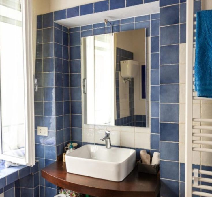 Ein einfaches Badezimmer mit blauen Fliesen, einem klassischen Waschbecken und einem großen Spiegel für gutes Licht.