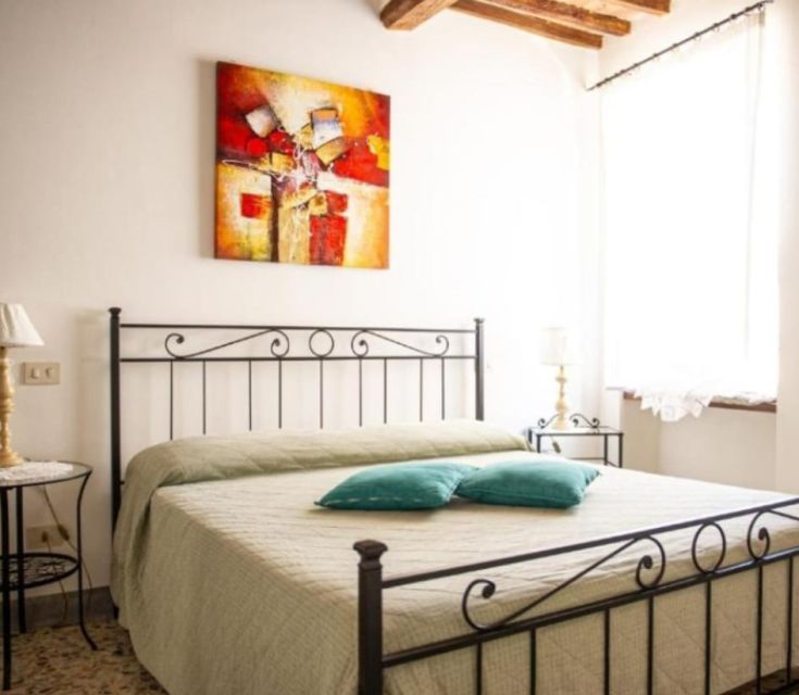 Ein Schlafzimmer mit einem Eisenbett und einem Fenster, das natürliches Licht hereinlässt. Ein Gemälde hängt an der Wand.