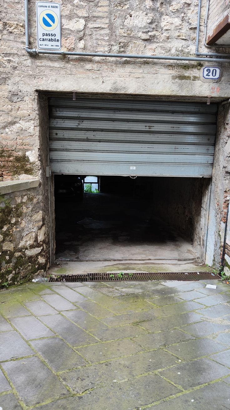 L'entrée d'un garage en pierre grise avec ouverture motorisée, ombragée et avec un sol mouillé.