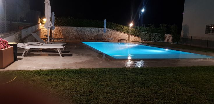 A lit swimming pool at night in a farmhouse, with sunbeds and umbrellas arranged around for relaxation.