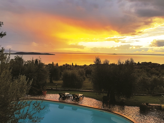 Panorama sul Lago Trasimeno al tramonto, circondato da ulivi e una piscina.