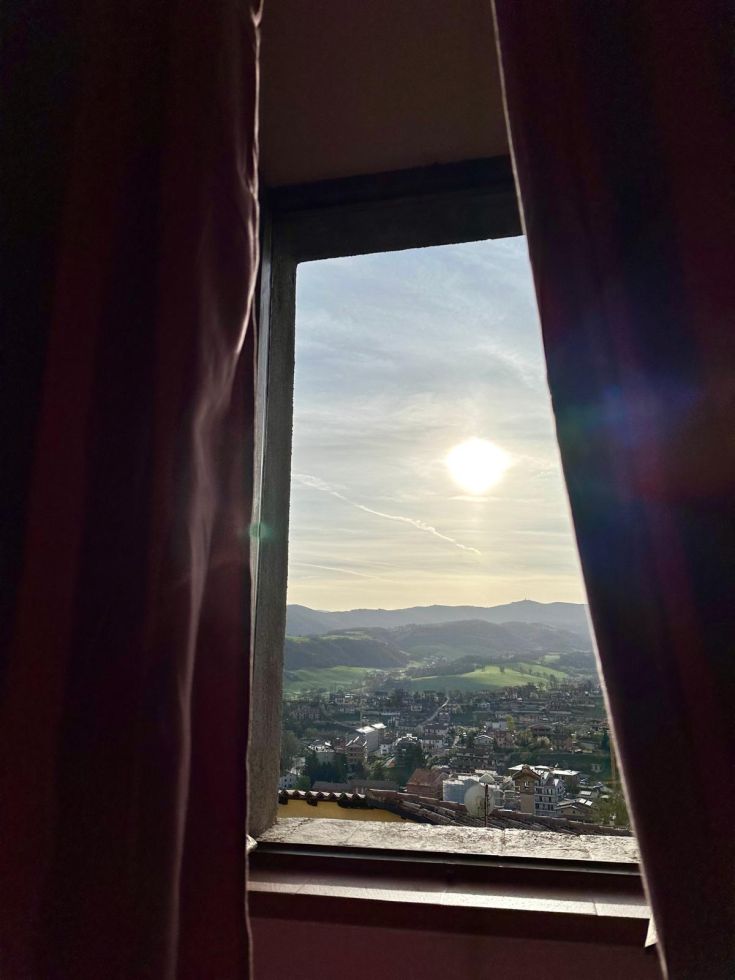 Scena di un paesaggio montano osservata da una finestra di un edificio storico, con il sole che sorge in lontananza.